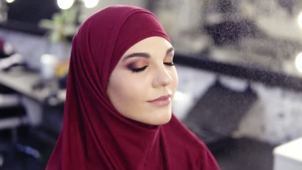 Hermosa chica joven con ojos de avellana y hiyab púrpura en su cabeza ha maquillaje niebla fijación rociado en su cara mientras recibe toques finales de impecable maquillaje — Vídeos de Stock