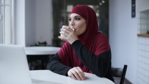 Jovem atraente tímida fêmea em hijab bebendo cappuccino em algum café minimalista. Imagens internas — Vídeo de Stock