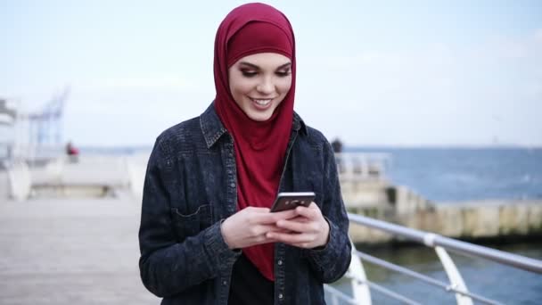 Muchacha joven atractiva con hijab en la cabeza está sonriendo mientras le envía mensajes a alguien y desplazando algo en su teléfono inteligente. Imágenes en cámara lenta al aire libre — Vídeos de Stock