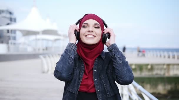 Slow motion footage of a young attractive girl with hijab on her head putting headphones on, smiling and enjoying the music while walking near sea side — Stock Video