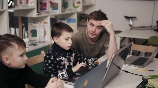 Joven profesor attactive en sus 20 años explicando algo a dos niños pequeños, enseñándoles cómo utilizar el ordenador portátil. Clase de programación. Proceso educativo . — Vídeos de Stock