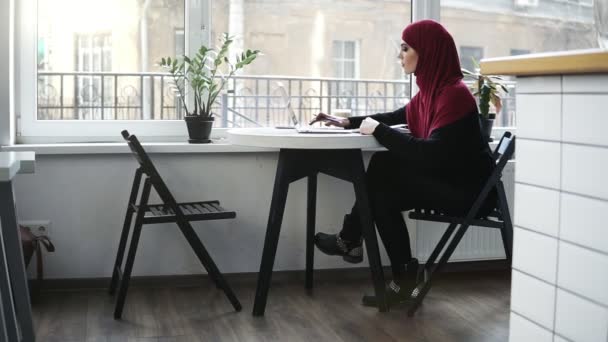 Hermosa chica musulmana usando hijab está buscando algo en Internet en su computadora portátil y escribe algo mientras está sentado en un lugar de luz minimalista en el interior — Vídeo de stock