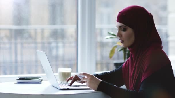 Chica musulmana atractiva está escribiendo algo de información de una tarjeta de visita en el teclado del ordenador portátil. Imágenes en interiores — Vídeos de Stock