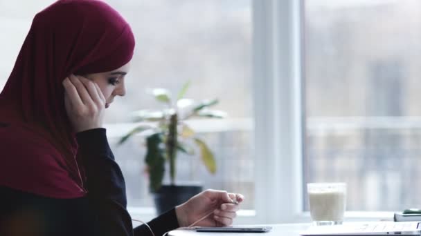 Una preciosa mujer árabe con hiyab se pone auriculares en los oídos y comienza a escuchar su música favorita. Imágenes en interiores — Vídeos de Stock