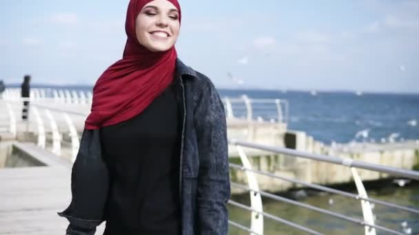 Atractiva chica musulmana con hiyab está sonriendo y yendo y viniendo, disfrutando de su paseo junto al mar. Actividades de tiempo libre. Imágenes al aire libre — Vídeos de Stock