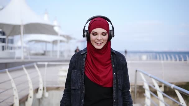 Moderne moslim meisje met hijab die betrekking hebben op haar hoofd zet koptelefoon op en begint te lopen ergens van genieten en dansen op de muziek — Stockvideo