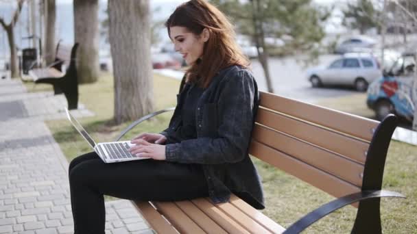 Atractiva hembra de unos 20 años con un atuendo urbano hipster sentada en el banco y escribiendo algo en el teclado de su portátil — Vídeos de Stock