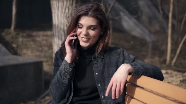 Красивая юная брюнетка в миниатюрном городском костюме сидит на скамейке в парке и разговаривает с кем-то по телефону — стоковое видео