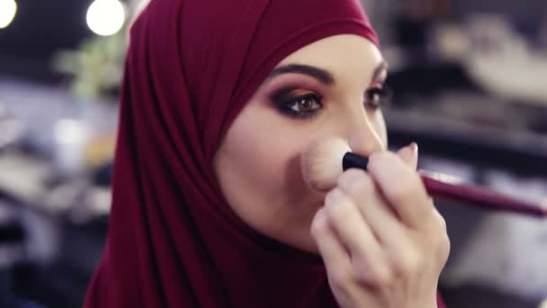 Bedövande vacker flicka med traditionella arabiska Lila chiffong hijab som täcker hennes huvud har oigenkännlig gör upp konstnärer hand tillämpa lite Rouge på kinderna. — Stockvideo