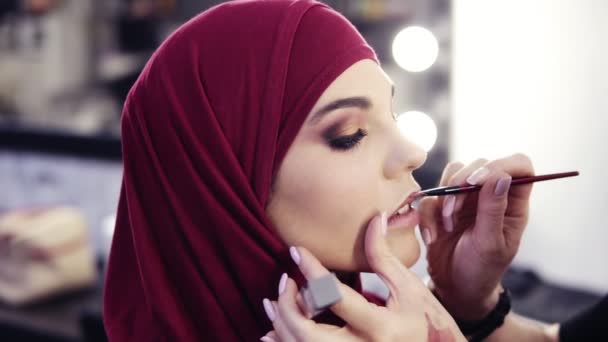 Невероятно красивая девушка с лесным орехом глаза и фиолетовый хиджаб на голове имеет неузнаваемый макияж руки художников аккуратно нанесения помады со специальной кистью — стоковое видео