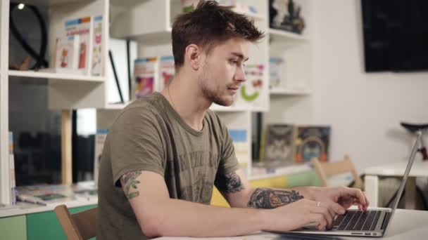 Professor programador jovem atraente com várias tatuagens em seus braços está murmurando algo enquanto digita no teclado do laptop . — Vídeo de Stock