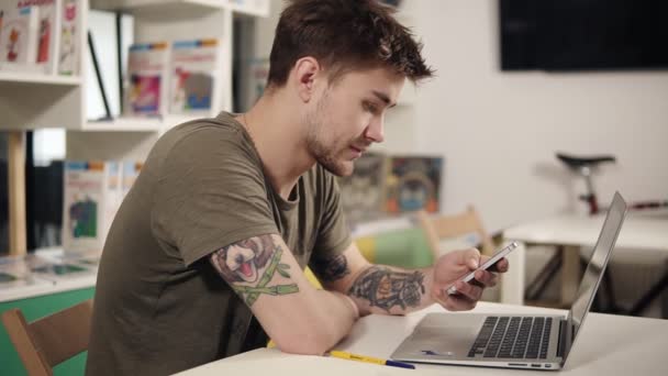 Aantrekkelijke programmeur met tattooes op zijn armen iets op zijn smartphone scrollen zittend in een klaslokaal naast laptop. — Stockvideo