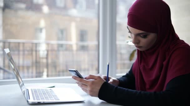 Menina jovem supostamente muçulmano com hijab roxo na cabeça escreve algo para baixo de seu smartphone, sentado em um espaço interior claro com janelas de vidro no fundo — Vídeo de Stock