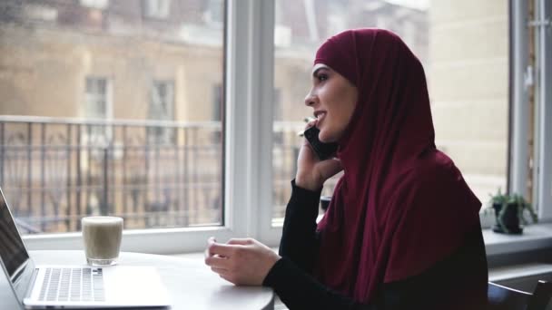 Gadis muda yang menarik dari kebangsaan yang tidak dikenal dengan hijab di kepalanya sedang berbicara dengan seseorang di telepon sementara dia memiliki laptop dan segelas cappuccino berbaring di depannya — Stok Video
