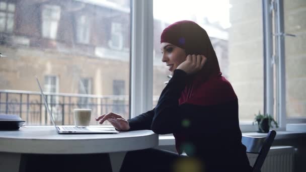 Aantrekkelijke moslim meisje met hijab die betrekking hebben op haar hoofd is op zoek en glimlachend op iets op haar laptop scherm. Binnenshuis slow motion beelden — Stockvideo