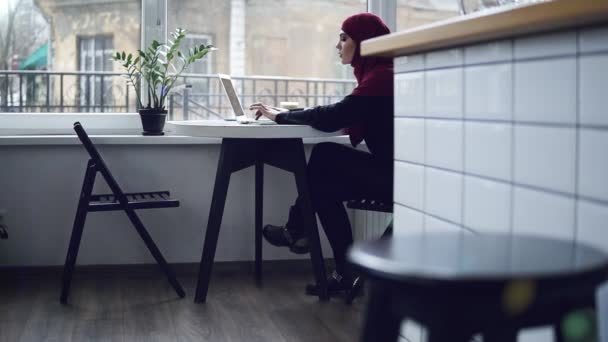 Hermosa chica musulmana con hijab en la cabeza, sentado cerca de la ventana, está escribiendo algo en el teclado de su computadora portátil — Vídeos de Stock