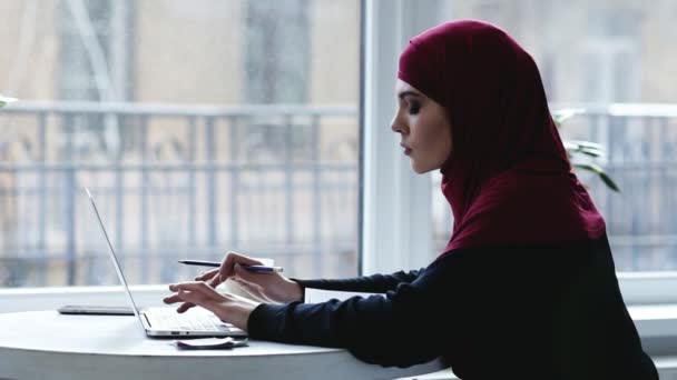 Indoors footage of beautiful muslim girl with hijab on her head typing something and then writing something down in her notebook — Stock Video