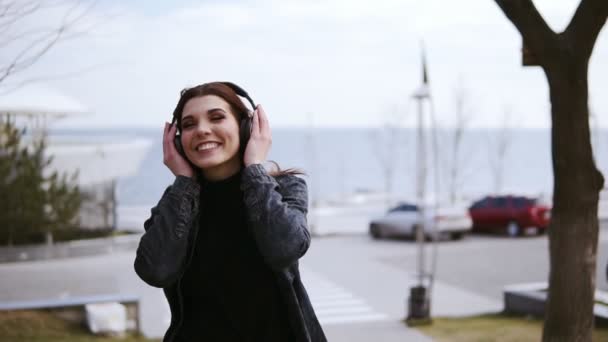 Menina morena jovem atraente com fones de ouvido sem fio preto está andando em algum lugar, ouvindo e cantando música. Desfrutando de juventude e tempo livre. Filmagem ao ar livre — Vídeo de Stock