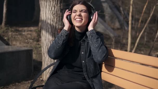 Schöne attraktive junge Mädchen in schwarzen Jeansjacken sitzt auf der Bank im Park und genießt Musik hören in ihren schwarzen drahtlosen Kopfhörern — Stockvideo