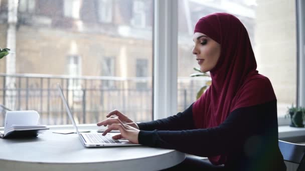 Unga attraktiva muslimsk flicka med hijab som täcker hennes huvud är att skriva något på sin laptop när man sitter i vissa förment Co arbetsrymden med glasfönster på bakgrunden — Stockvideo