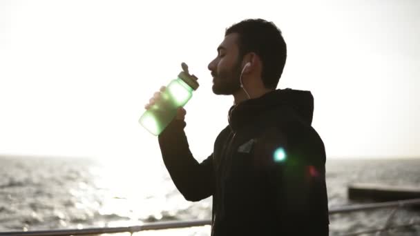 Joven attarctive masculino con auriculares en los oídos beber agua de plástico botella de fitness después del entrenamiento. Deporte, fitness, bienestar juvenil . — Vídeo de stock
