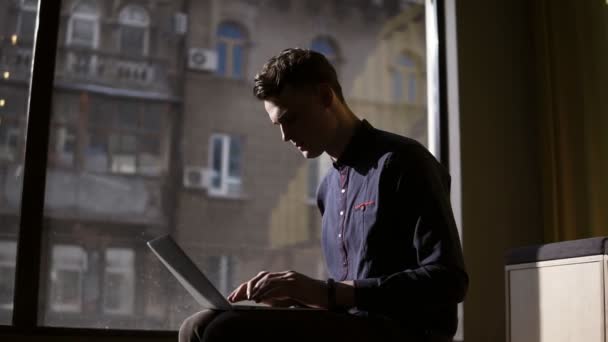 Atractivo joven sexy sentado cerca de ventana panorámica escribiendo algo en su computadora portátil. Proceso de trabajo . — Vídeos de Stock