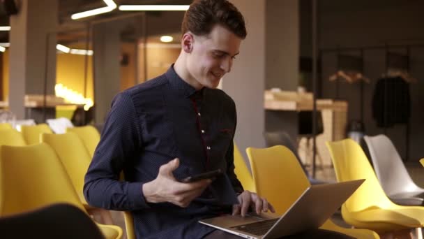 Jeune homme attrayant dans une chemise mignonne souriant tout en tenant smartphone et en tapant quelque chose sur son ordinateur portable en même temps . — Video