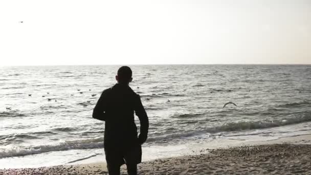 Молодой спортивный мужчина с кавказским внешним видом в черной спортивной одежде бежит вдоль побережья с подключенными наушниками. Красивый живописный вид — стоковое видео
