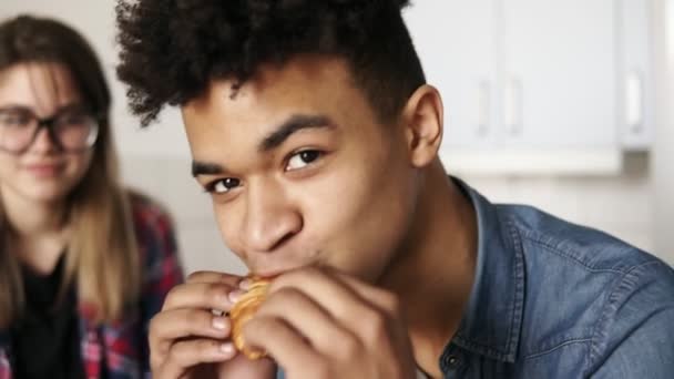 Närbild bilder av en stilig mulatt kille i 20-årsåldern med uttrycksfulla djupa mörka bruna ögon tittar på kameran, äter croissant, med sin flickvän som sitter i bakgrunden. — Stockvideo