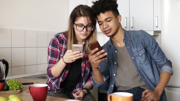 Atractivo chico y una chica tanto en sus 20 años desplazando sus teléfonos inteligentes, el intercambio de información mientras se sienta en una cómoda cocina espaciosa . — Vídeo de stock