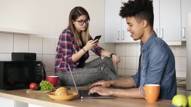 Jovem casal feliz de hipsters sentados na cozinha, ambos usando alguns dispositivos como laptop, smartphone, não falando uns com os outros . — Vídeo de Stock