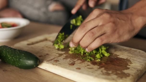 Close-up van slow-motion beelden van een jonge mulat kerel snijden sla voor salade, maken van de lunch met zijn vriendin wie naast hem op het oppervlak van de tabel keuken zit. — Stockvideo