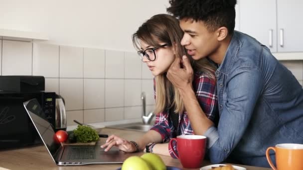 Caucasienne fille à la recherche et son petit ami mulâtre à la fois dans la vingtaine en utilisant un ordinateur portable, surfer sur le web dans la cuisine à la maison. Il l'embrasse tendrement par le dos . — Video