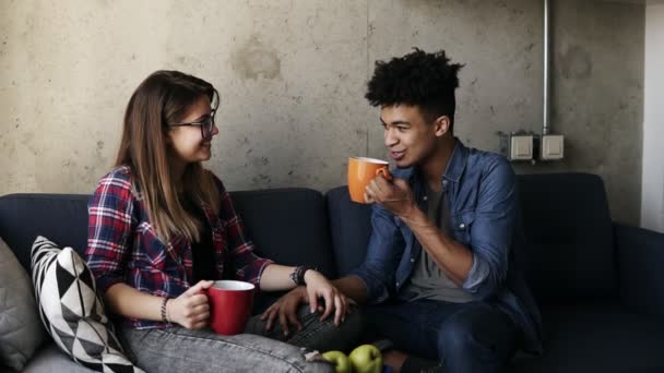 Young caucasian girl and handsome mulatto guy both wearing urban outfits, sitting on the couch with cups of tea, talking about life, enjoying spending time together. — Stock Video