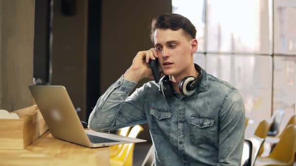 Привлекательный мужчина 20 лет в джинсовой рубашке с беспроводными наушниками на шее и ноутбуком рядом с ним, разговаривает с кем-то по телефону. Открытое коворкинг пространство. В помещении . — стоковое видео