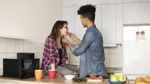 Deux jeunes profitent de leur temps ensemble, préparant le déjeuner dans une cuisine confortable. Vivre ensemble. Mulatto gars est nourrir tranche de tomate à son attrayant caucasien copine . — Video