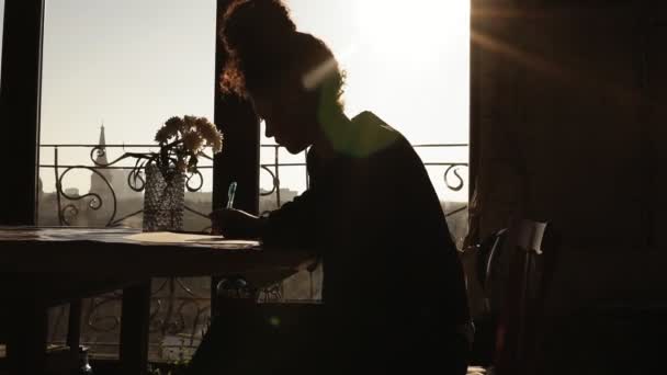 Sombreado por el sol, figura de un joven artista creativo dibujando algo, sentado junto a la mesa con hermosas flores en ella. Artesanía inspiradora . — Vídeo de stock