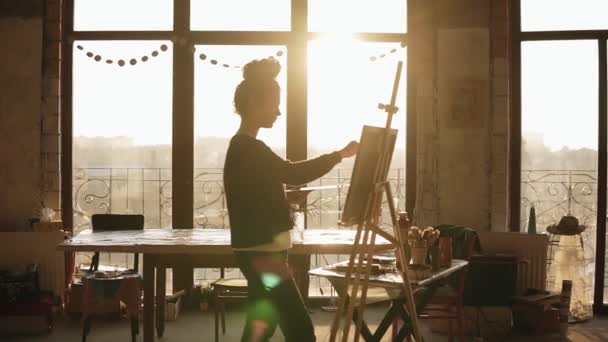 Fully concentrated sophisticated female artist in her 20s is drawing picture on easel. The sun behind lits up the art studio and turns it into inspiring environment of creativity. — Stock Video