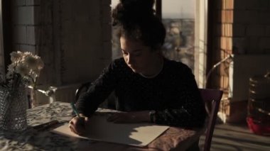 Kıvırcık saçlı bir topuz çekti genç sevimli kadın sanatçı, masaya oturan ve tam bir kalemle çizim oluşturma işlemi üzerinde yoğunlaştı. Resim sanatı, ilham.