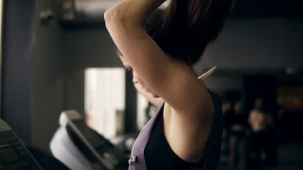 Slowmotion footage of a sportive girl with long brunette hair putting headphones on and choosing favourite music track before starting the session on treadmill. — Stock Video