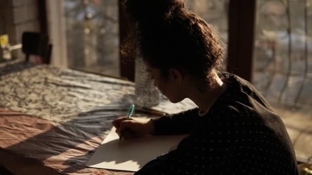 Linda artista feminina com seu cabelo encaracolado puxado em um pão sentado à mesa perto da janela panorâmica em um estúdio de arte com interior minimalista e desenho com uma caneta no papel — Vídeo de Stock