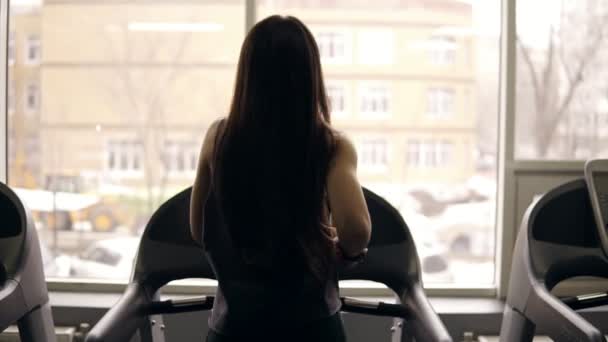 Backside footage in slow motion of a sportive girl with long brunette hair running on a treadmill. Indoors. — Stock Video