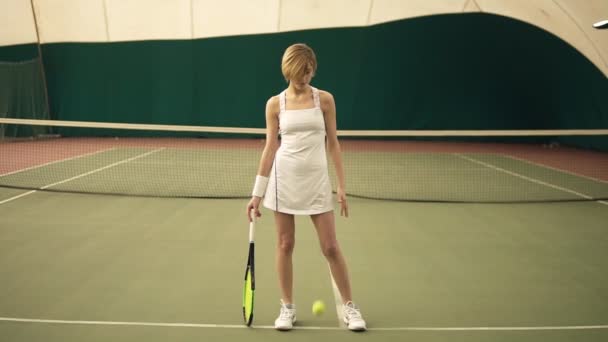 Ritratto full size di una giovane attraente donna sportiva con capelli corti biondi che indossa abbigliamento sportivo bianco, giocando con la palla nel campo da tennis coperto — Video Stock