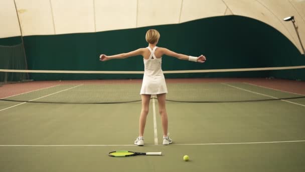 Rückseite Zeitlupenaufnahmen einer jungen, sportlichen Tennisspielerin mit kurzen blonden Haaren, die weiße Sportbekleidung beim Aufwärmen trägt und die Muskeln vor dem Spiel dehnt — Stockvideo