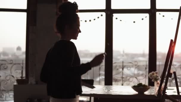 Giovane artista femminile riccia carino ballare in studio d'arte poi finisce le linee sul suo dipinto, con lo sfondo di un bel tramonto . — Video Stock