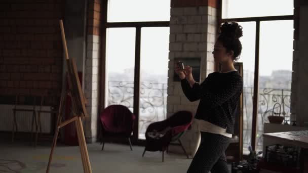 Krullend meisje trots op zichzelf en haar werk nemen foto op haar smartphone van het doek met de schilderkunst trok zij. — Stockvideo