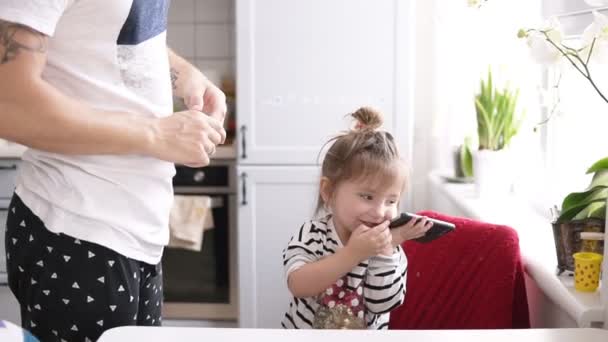 Мила маленька дівчинка розмовляє по телефону, тато стоїть поруч на кухні — стокове відео