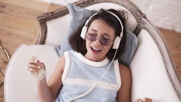 Élégante fille hipster dans la vingtaine portant des écouteurs, des lunettes et des gants de motard en cuir punk, couchée sur le canapé, chantant et dansant sur la musique — Video