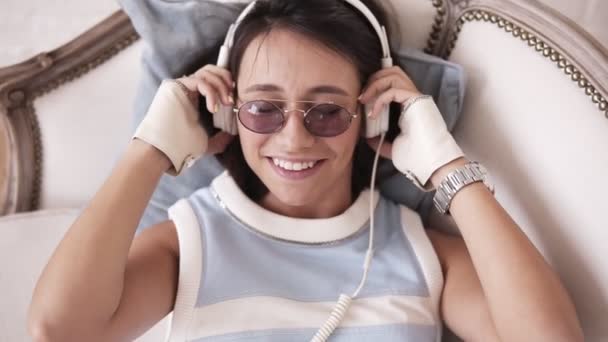 Muchacha alegre atractiva con auriculares y gafas sonriendo y bailando a la música mientras está acostado en el sofá — Vídeos de Stock