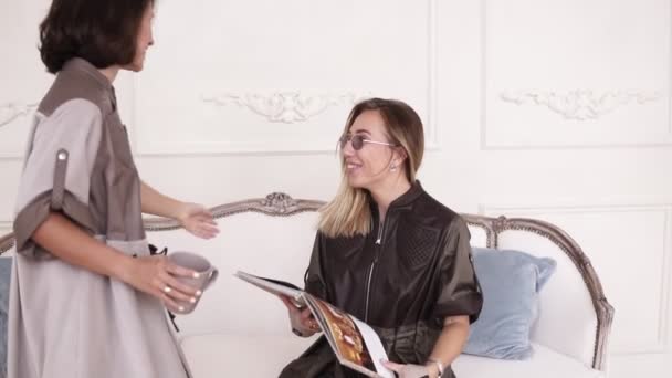 Deux copines élégantes se rencontrent dans un espace intérieur confortable, avec des murs blancs sur le fond. Les filles s'embrassent sur les joues. Fille parler — Video